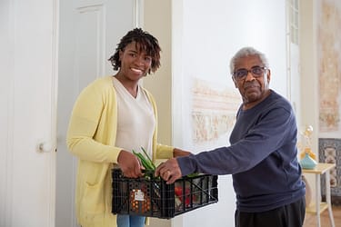 a-man-and-woman-holding-a-plastic-crater-7551593 Smiling woman assists a senior man with food delivery indoors, promoting community support and care.