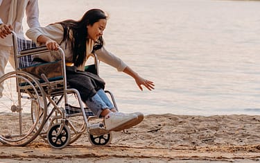 woman-reaching-for-the-sand-while-sitting-on-the-wheelchair-7699053 Asian woman in a wheelchair enjoying time by the ocean on a sandy beach, reaching out.