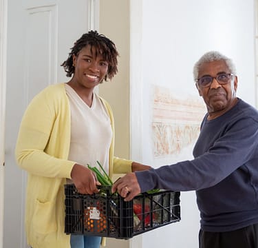 a-man-and-woman-holding-a-plastic-crater-7551593 Smiling woman assists a senior man with food delivery indoors, promoting community support and care.