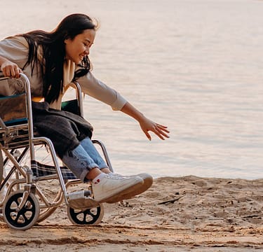 woman-reaching-for-the-sand-while-sitting-on-the-wheelchair-7699053 Asian woman in a wheelchair enjoying time by the ocean on a sandy beach, reaching out.