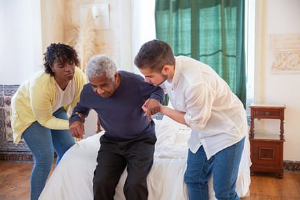 a-man-and-a-woman-assisting-an-elderly-man-in-standing-7551686 An elderly man receiving assistance from caregivers in a cozy home environment.