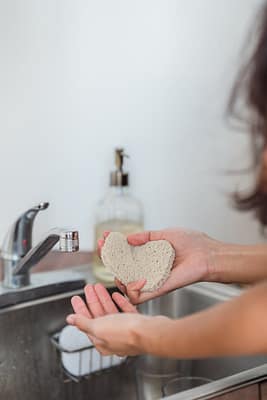 a-person-holding-a-scrub-9475295 Hands holding a heart-shaped sponge by a kitchen sink, suggesting cleaning and hygiene.